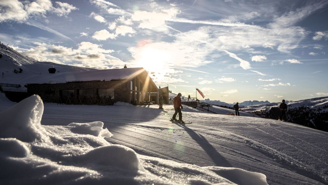 Ein Skifahrer auf einer schneebedeckten Piste vor einer Berghütte, wo die tief stehende Sonne lange Schatten erzeugt.