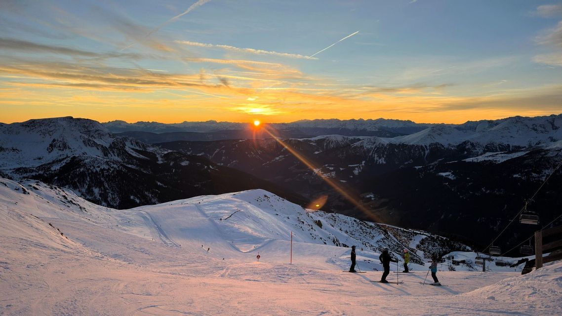 Eine WeRoad-Gruppe von Skifahrern pausiert auf einem verschneiten Berghang und genießt den Sonnenuntergang hinter fernen Gipfeln.