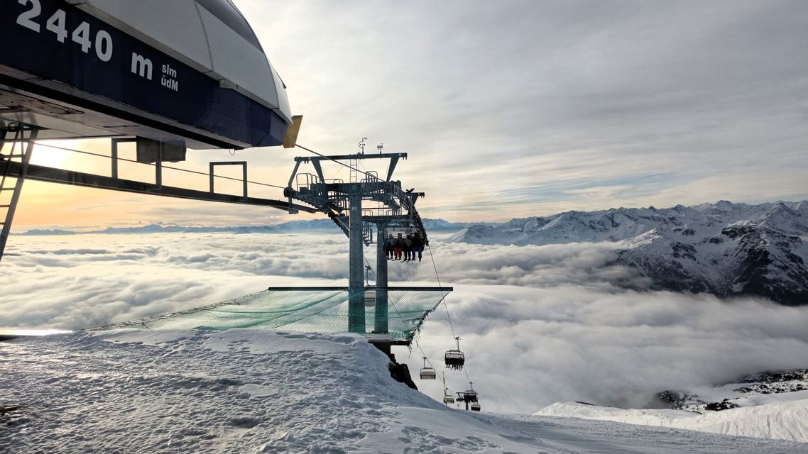 Eine WeRoad Gruppenreise im Skilift, der von einer verschneiten Bergstation über einem Wolkenmeer abfährt.