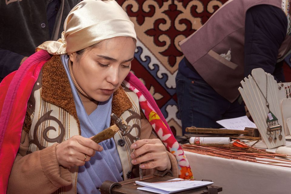 Una donna con un foulard si concentra mentre modella un pezzo di metallo con un piccolo martello su un tavolo da artigianato.