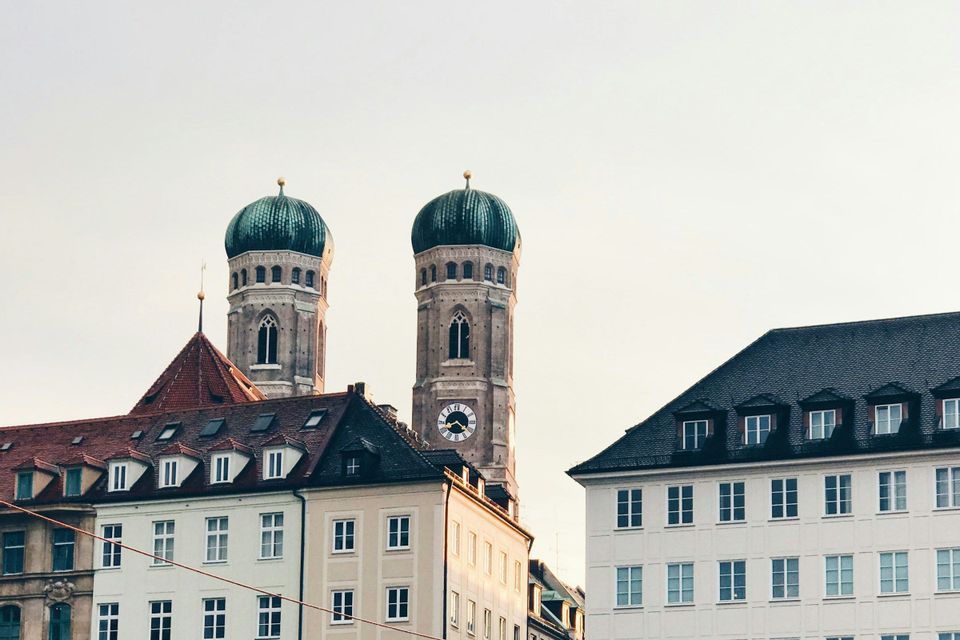 Le torri gemelle di una cattedrale con cupole verdi e un orologio si ergono sopra i tetti degli edifici della città contro un cielo pallido.
