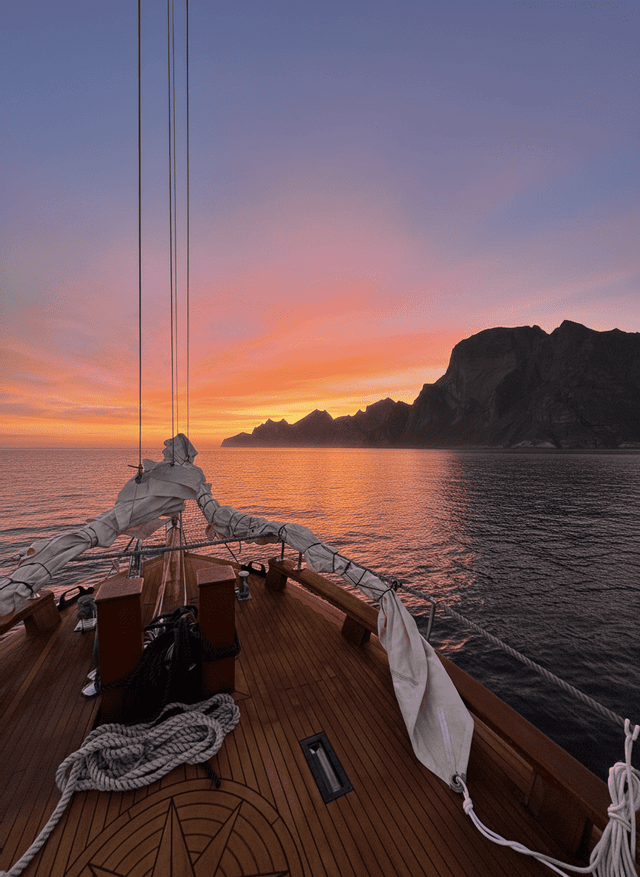 La vista dal ponte in legno di una barca a vela che si affaccia su una costa montuosa durante un tramonto colorato.
