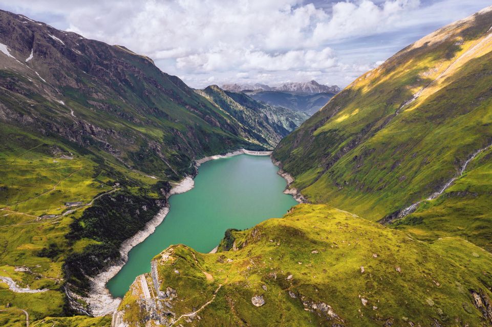 Luftaufnahme eines türkisfarbenen Alpensees in einem tiefen Tal zwischen steilen, grünen Bergen unter bewölktem Himmel.