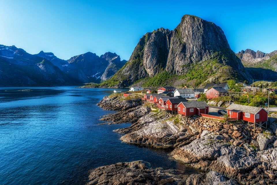 Ein Küstendorf mit roten Holzhäusern, an einer felsigen Küste neben einem Fjord gelegen, mit steilen Bergen im Hintergrund unter klarem Himmel.