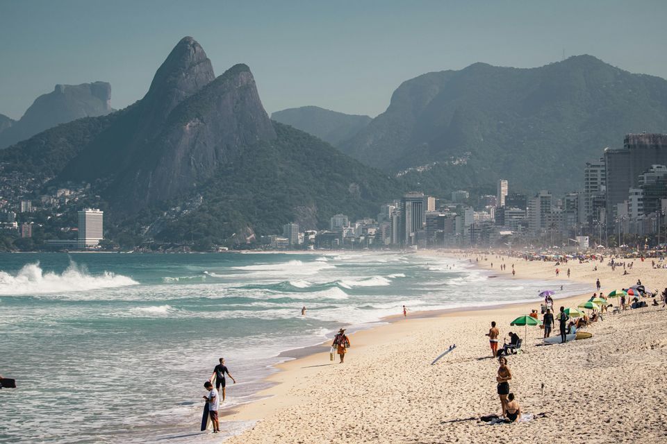Un'ampia veduta di una spiaggia urbana affollata, con persone sulla sabbia, sullo sfondo uno skyline cittadino e grandi montagne alberate.