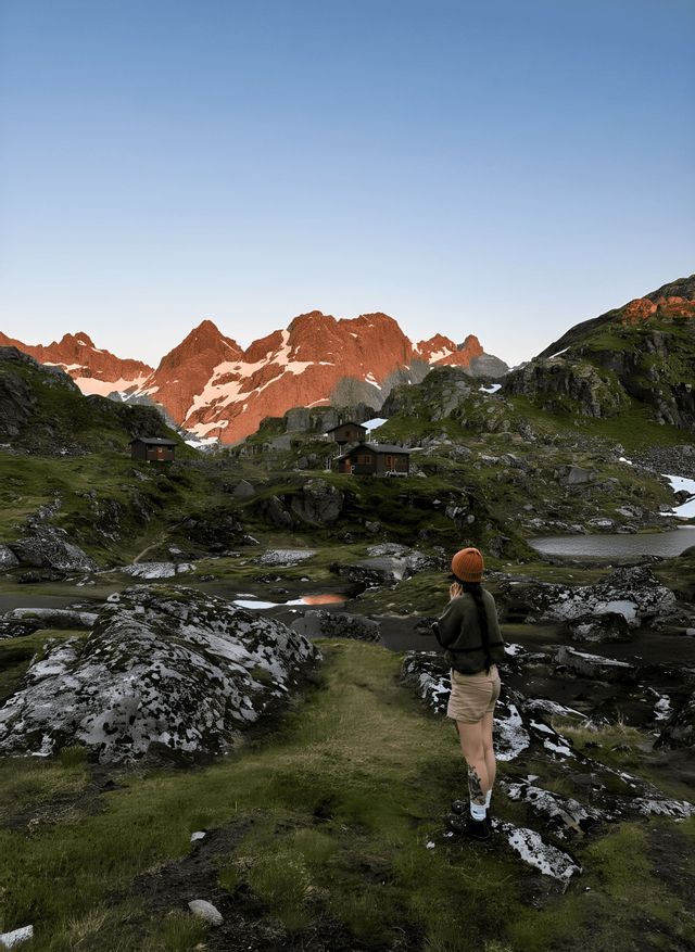 Eine Person steht auf einem grünen, felsigen Wanderweg und blickt auf ein Tal mit Holzhütten, einem See und sonnenbeschienenen, schneebedeckten Bergen.