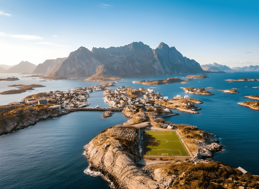 Veduta aerea di un villaggio costiero e di un campo da calcio su isole rocciose, con grandi montagne che si ergono dal mare sullo sfondo.