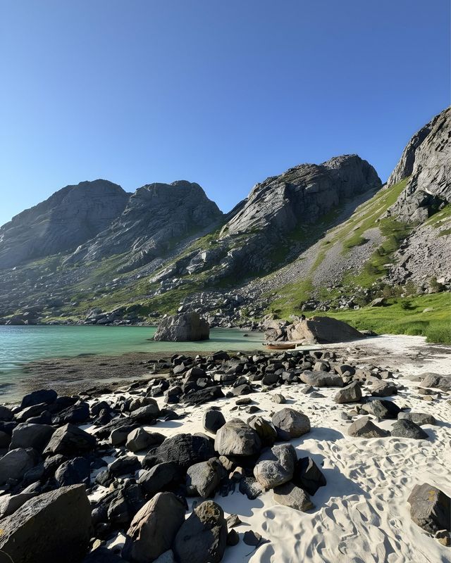 Ein sandiger und felsiger Strand grenzt an ruhiges, türkisfarbenes Wasser am Fuße großer, grüner Berge unter einem klaren blauen Himmel.