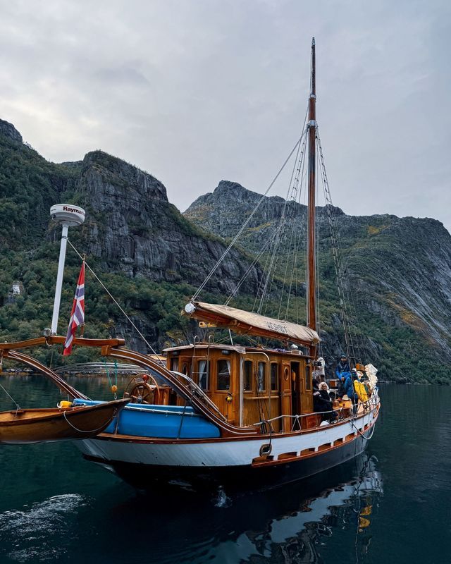Un viaggio di gruppo WeRoad su una barca a vela in legno naviga lungo un fiordo con ripide montagne rocciose sullo sfondo.