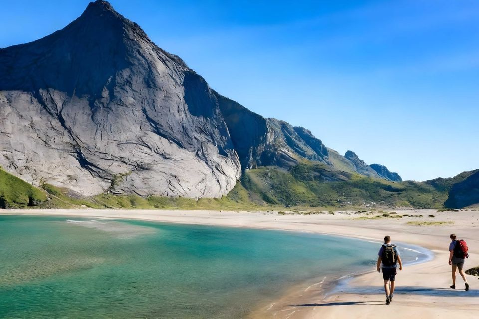 Zwei Wanderer mit Rucksäcken gehen an einem Sandstrand neben türkisfarbenem Wasser am Fuße eines hoch aufragenden Felsbergs.