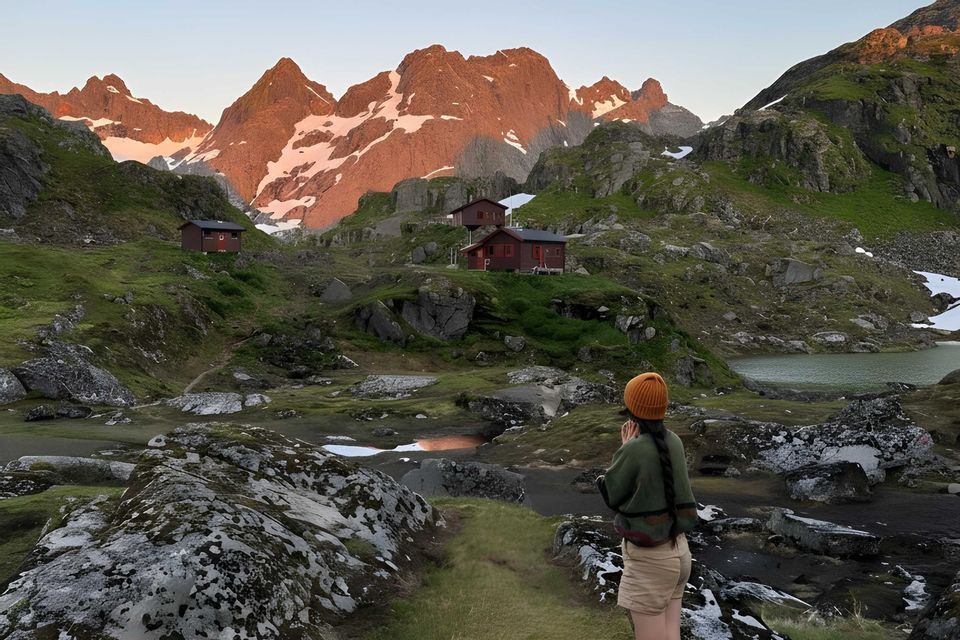 Una donna con un berretto giallo è su un sentiero che si affaccia su una valle verde con baite rosse, un lago e cime montuose innevate illuminate dal sole.