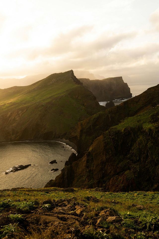 Scogliere scoscese e ricoperte di verde si protendono nell'oceano, circondando una baia tranquilla sotto la calda luce del sole.