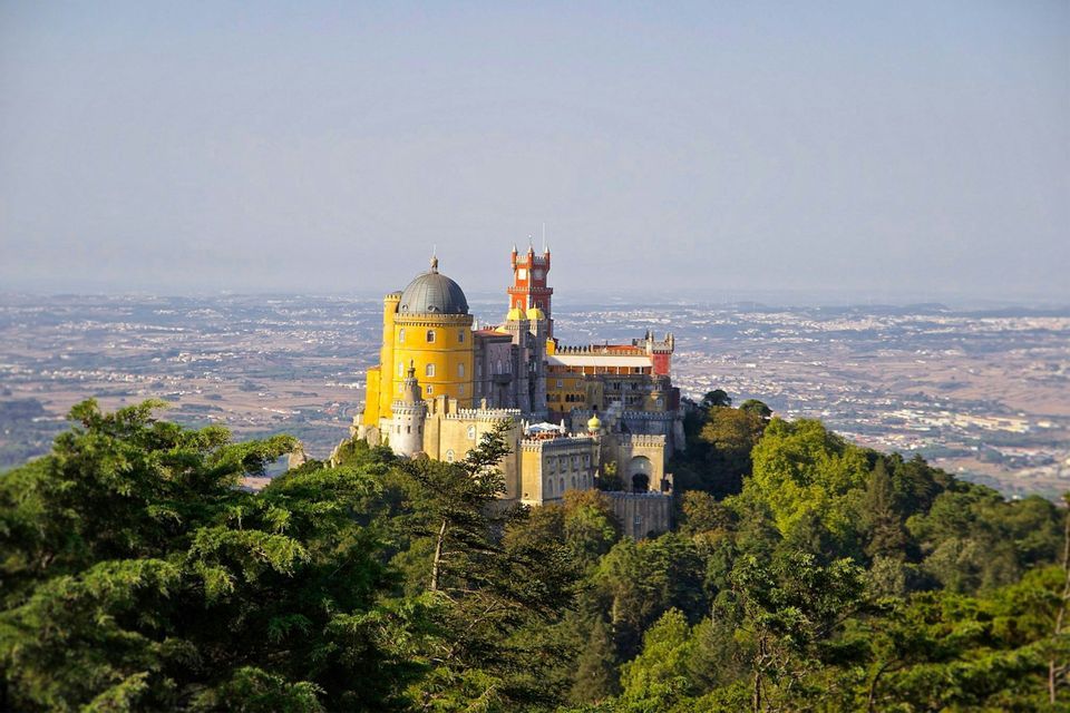 Un château coloré aux tours jaunes et rouges se dresse sur une colline boisée, dominant un vaste paysage.