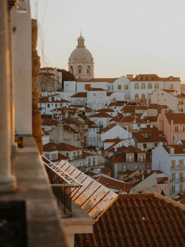 Una vista dall'alto di una città con edifici bianchi e tetti di tegole rosse, con una grande cupola in pietra visibile in lontananza al tramonto.