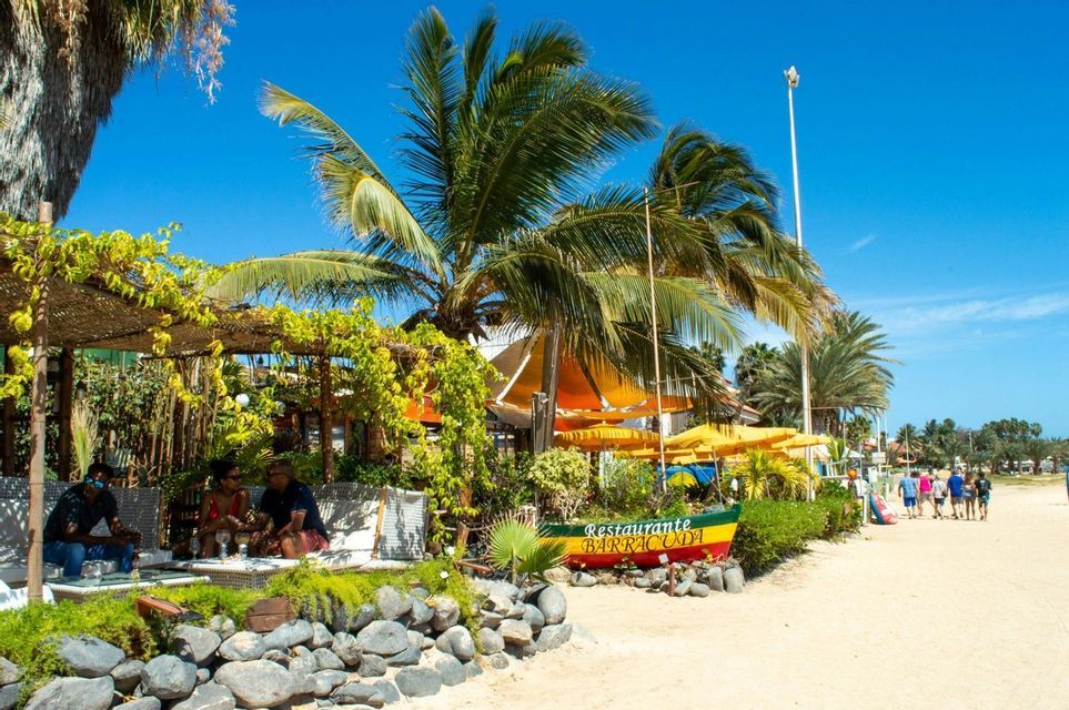 A WeRoad group trip walking along a sandy path next to a tropical beachside restaurant surrounded by palm trees.