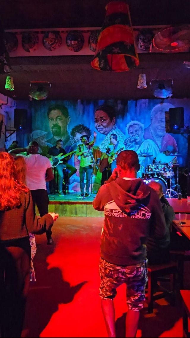 In einem schummrigen Club spielt eine Live-Band auf einer Bühne mit einem großen Wandgemälde im Hintergrund vor Publikum.