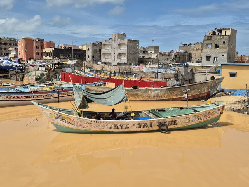 Una colorida barca de pesca con un toldo de lona flota en agua fangosa en un puerto abarrotado, con un paisaje urbano de fondo.