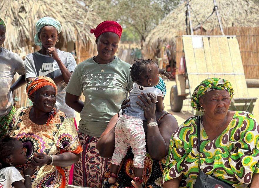Un grupo de mujeres y niños con ropa colorida y pañuelos en la cabeza se reunió al aire libre en un pueblo con cabañas con techo de paja.