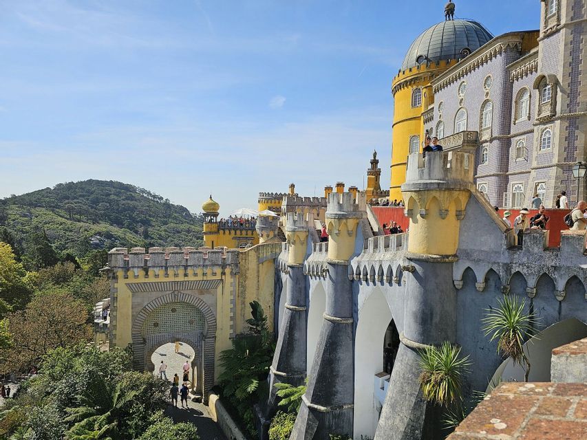 Un château jaune et bleu coloré, doté de tours et de remparts, avec des visiteurs sur ses chemins de ronde, sur fond de colline verdoyante et boisée.