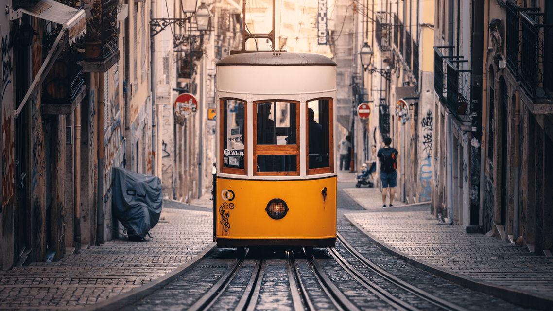 Un tram vintage giallo e bianco percorre i binari lungo una stretta strada acciottolata fiancheggiata da vecchi edifici.