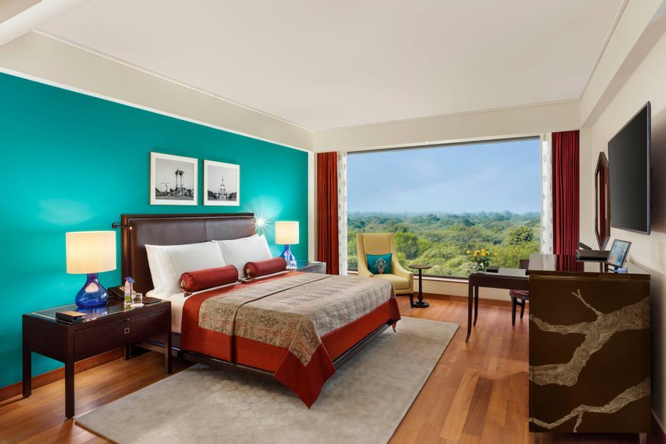 Una habitación de hotel ordenada con una cama doble junto a una pared turquesa y un gran ventanal con vistas a un frondoso bosque verde.
