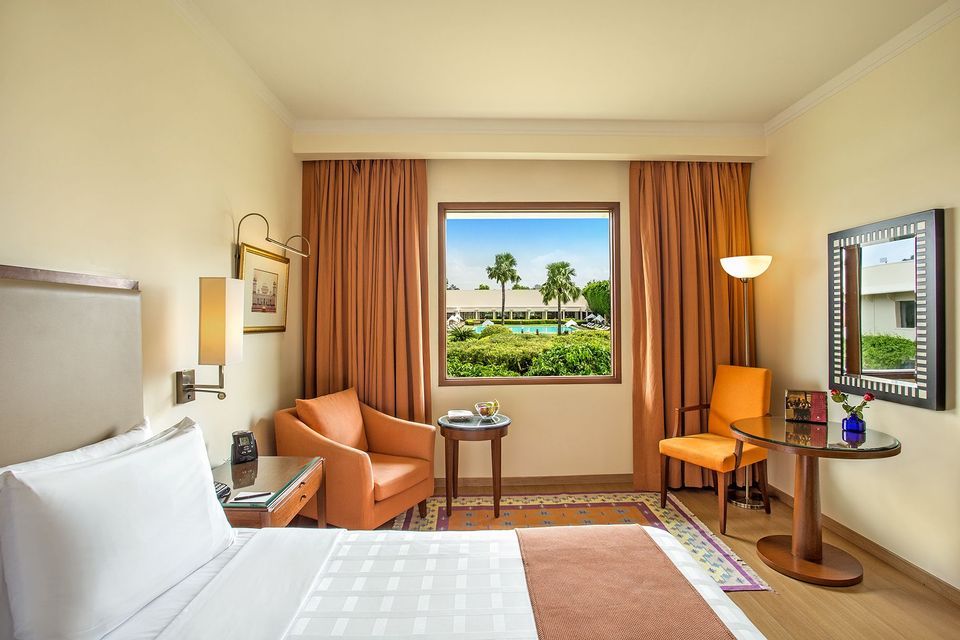 Una habitación de hotel con una cama bien hecha y sillas naranjas, que cuenta con un gran ventanal con vistas a la piscina del resort y palmeras.