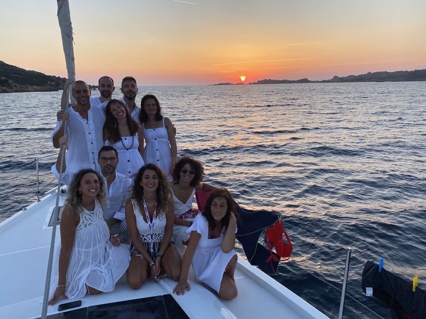 Un viaggio di gruppo WeRoad, tutti vestiti di bianco, in posa insieme sul ponte di una barca a vela al tramonto.