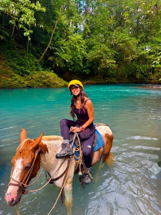 Una donna sorridente con un casco giallo cavalca un cavallo attraverso un fiume turchese in una lussureggiante foresta verde.