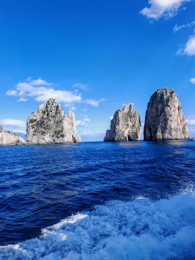 Grandi formazioni rocciose si ergono dal mare blu profondo sotto un cielo soleggiato, con la scia bianca di una barca in primo piano.