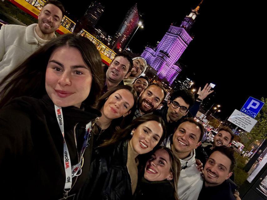 A WeRoad group trip smiles for a selfie on a city street at night, with an illuminated building and a tram in the background.