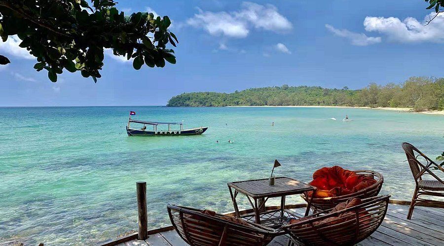Une table et des chaises sur une terrasse en bois surplombent une mer turquoise avec un bateau ancré et une plage tropicale lointaine.