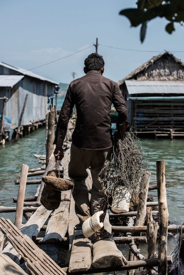 Un homme de dos à l'appareil photo transporte du matériel de pêche sur une jetée en bois dans un village sur pilotis.