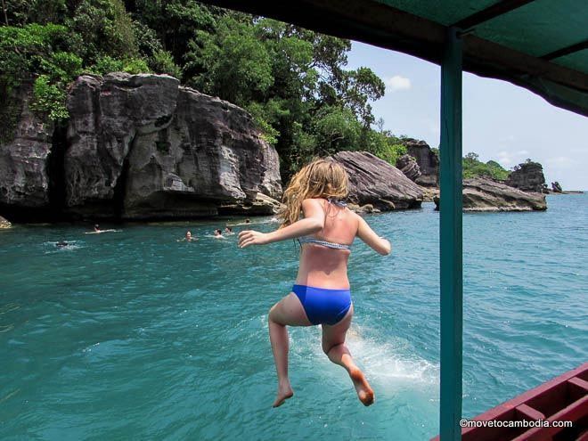 Une femme en bikini bleu saute d'un bateau dans l'eau turquoise, avec des falaises rocheuses et d'autres personnes qui nagent en arrière-plan.
