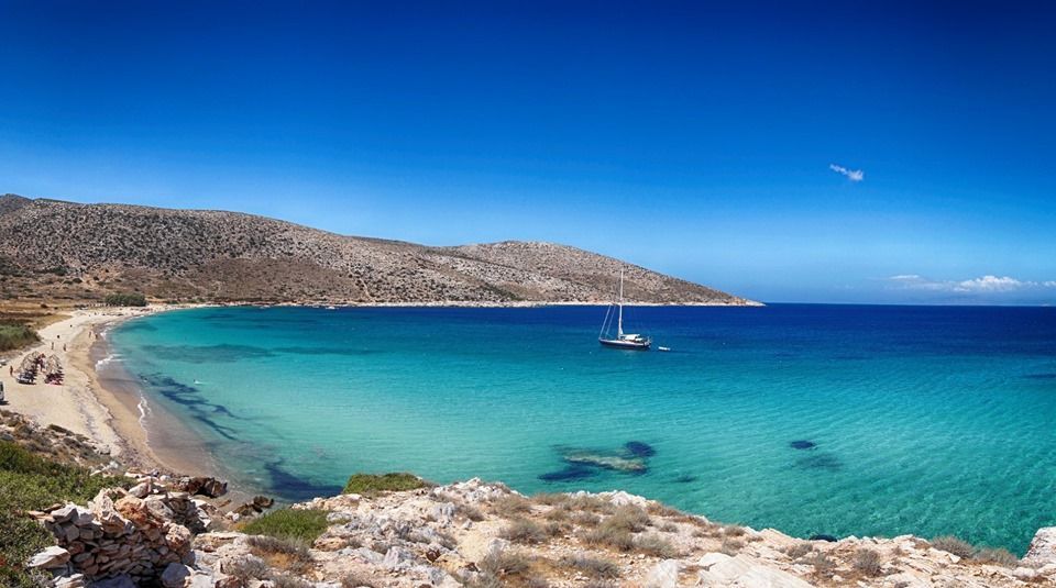 Una barca a vela ancorata in una baia con acqua limpida e turchese, una spiaggia di sabbia e colline rocciose sotto un cielo azzurro brillante.