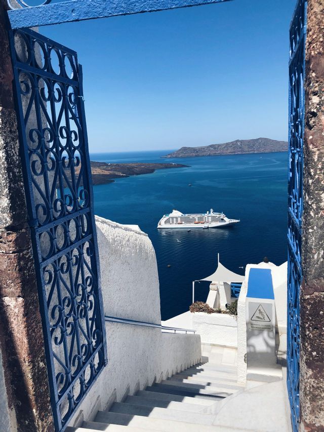 Incorniciata da due cancelli blu, una nave da crociera bianca solca il mare blu profondo, vista dalla cima di una scalinata imbiancata.