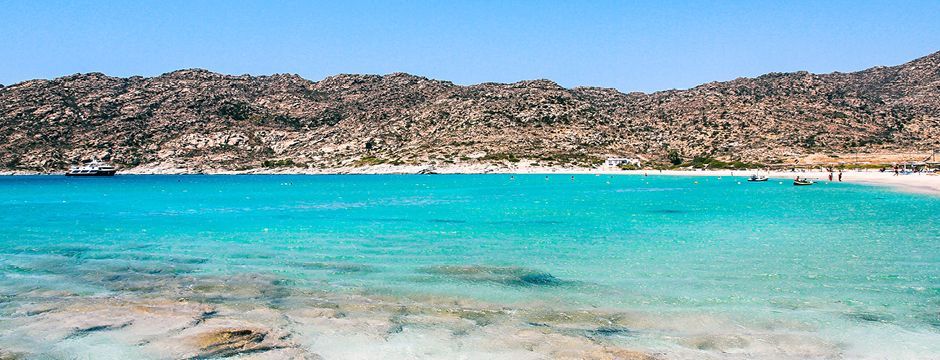 Vista panoramica di una baia con acqua cristallina color turchese, una spiaggia di sabbia e una catena montuosa rocciosa sotto un cielo azzurro splendente.
