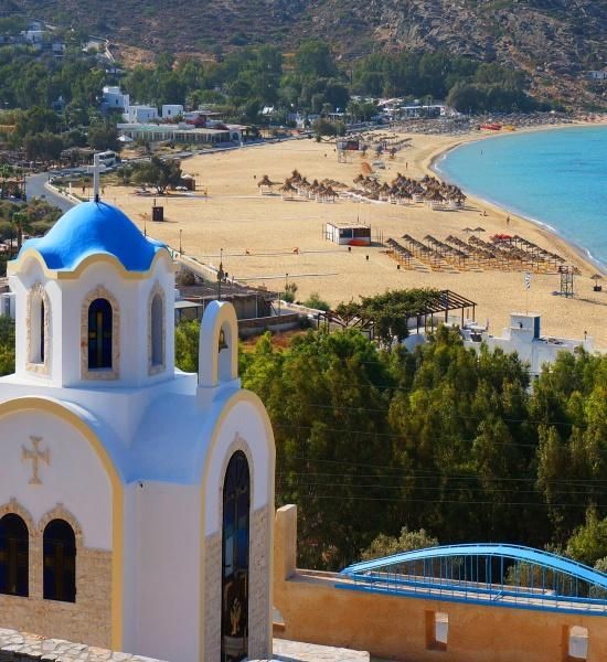 Una chiesa bianca con cupola blu si affaccia su un'ampia spiaggia sabbiosa con acqua turchese e file di ombrelloni.