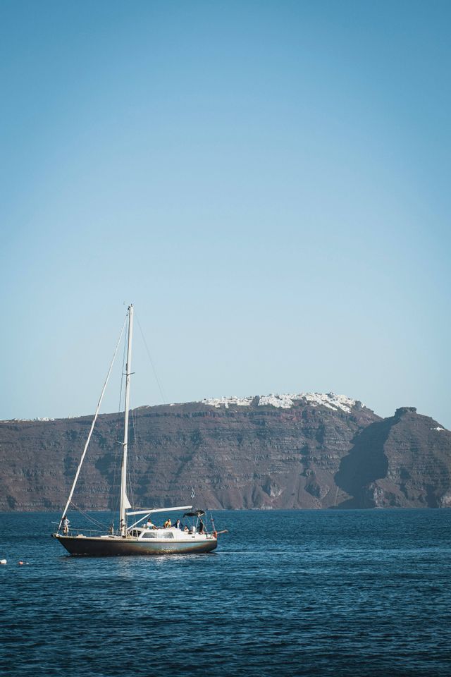 Una barca a vela con un gruppo WeRoad naviga sul mare blu di fronte a un'imponente scogliera con edifici bianchi in cima.