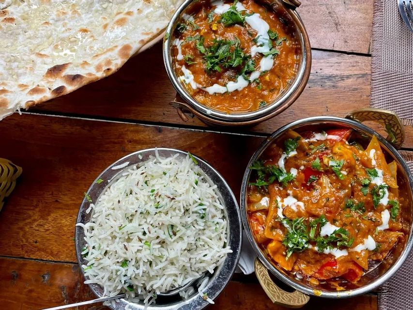 Una foto dall'alto di un pasto indiano con due ciotole di curry, una di riso speziato e pane naan su un tavolo di legno.