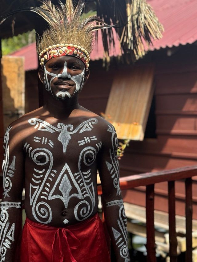 Un uomo con un copricapo di piume, il viso, il petto e le braccia ornati da un'intricata pittura tradizionale bianca.