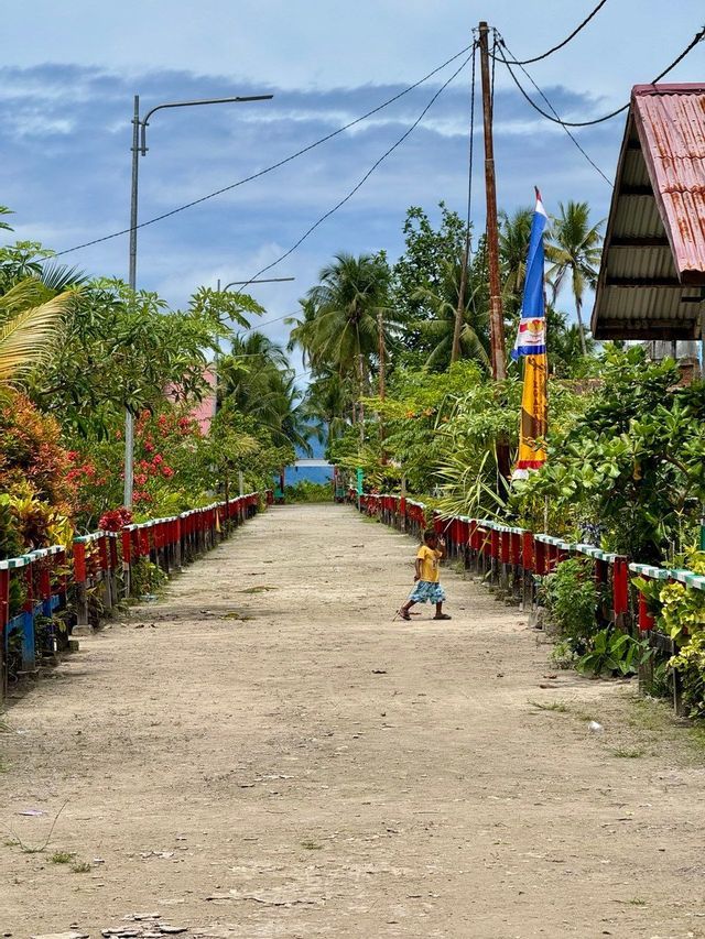 Un bambino cammina lungo un sentiero sterrato costeggiato da una recinzione colorata e rigogliose piante tropicali, verso il mare.
