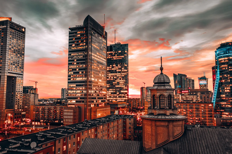 Uno skyline cittadino moderno con grattacieli illuminati ed edifici storici sullo sfondo di un drammatico tramonto arancione e nuvole scure.
