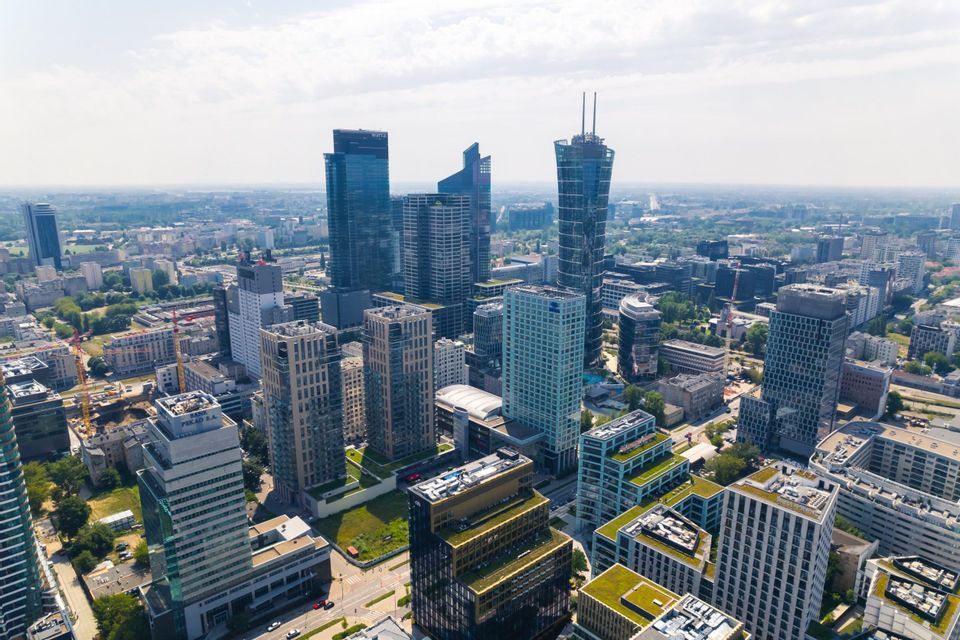 An aerial view of a modern city with glass skyscrapers and green-roofed buildings under a partly cloudy sky.