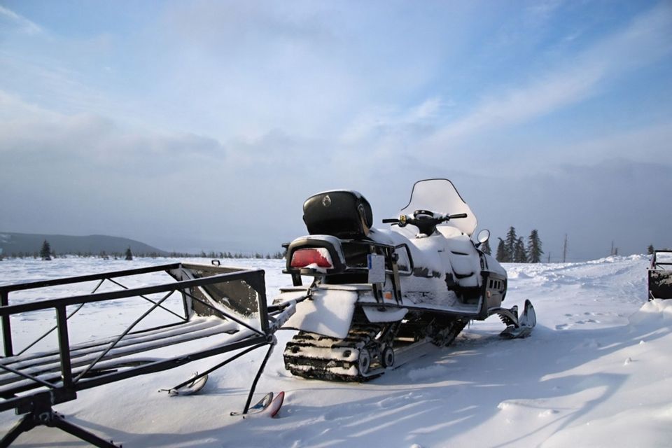 Ein schneebedecktes Schneemobil mit angehängtem Schlitten steht in einer weiten, verschneiten Landschaft unter einem bewölkten blauen Himmel.