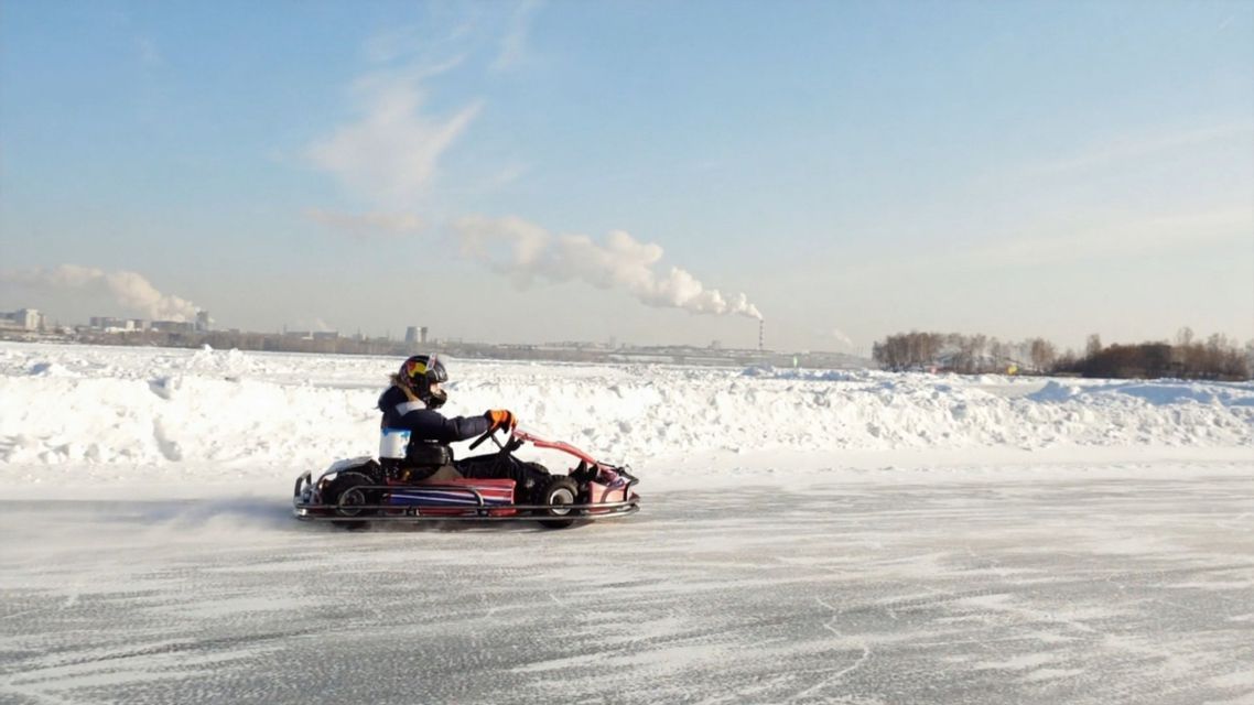 Eine Person mit Helm fährt ein Go-Kart auf einer gefrorenen, vereisten Strecke in einer verschneiten Landschaft mit einer Stadt in der Ferne.