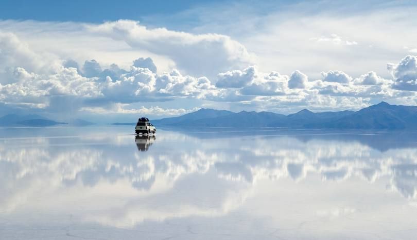 Un SUV bianco attraversa una vasta distesa salata, la sua superficie bagnata riflette il cielo nuvoloso e le montagne come uno specchio.