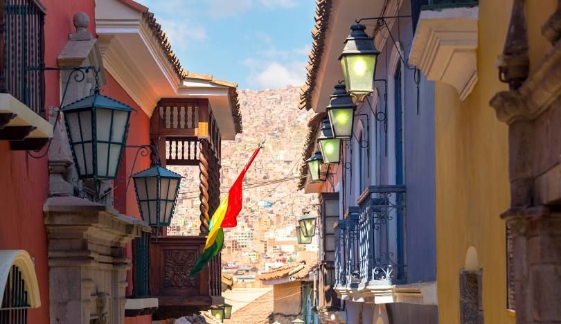 Uno scorcio di una stretta via coloniale con muri colorati, lanterne e una bandiera boliviana, che si affaccia su una città costruita su una collina.