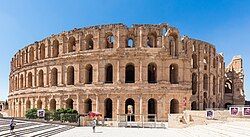 Un grand amphithéâtre romain en pierre, avec trois niveaux d'arches, se dresse sous un ciel bleu clair.