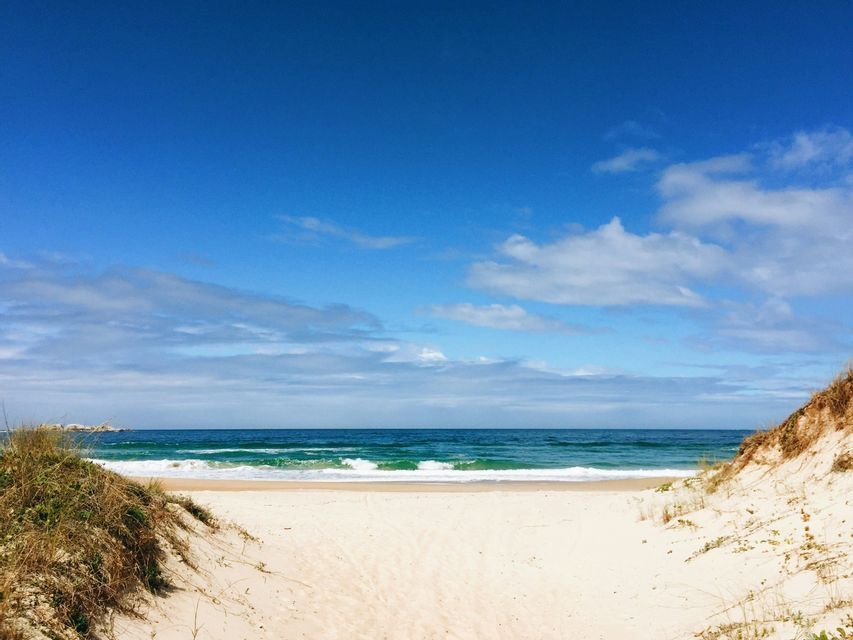 Un sentiero sabbioso tra due dune erbose si apre su una spiaggia con dolci onde oceaniche sotto un cielo blu con nuvole sparse.