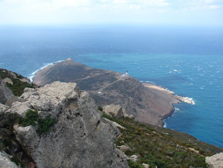 Vue d'une falaise rocheuse dominant une péninsule avec un phare blanc, entourée par la mer bleue.